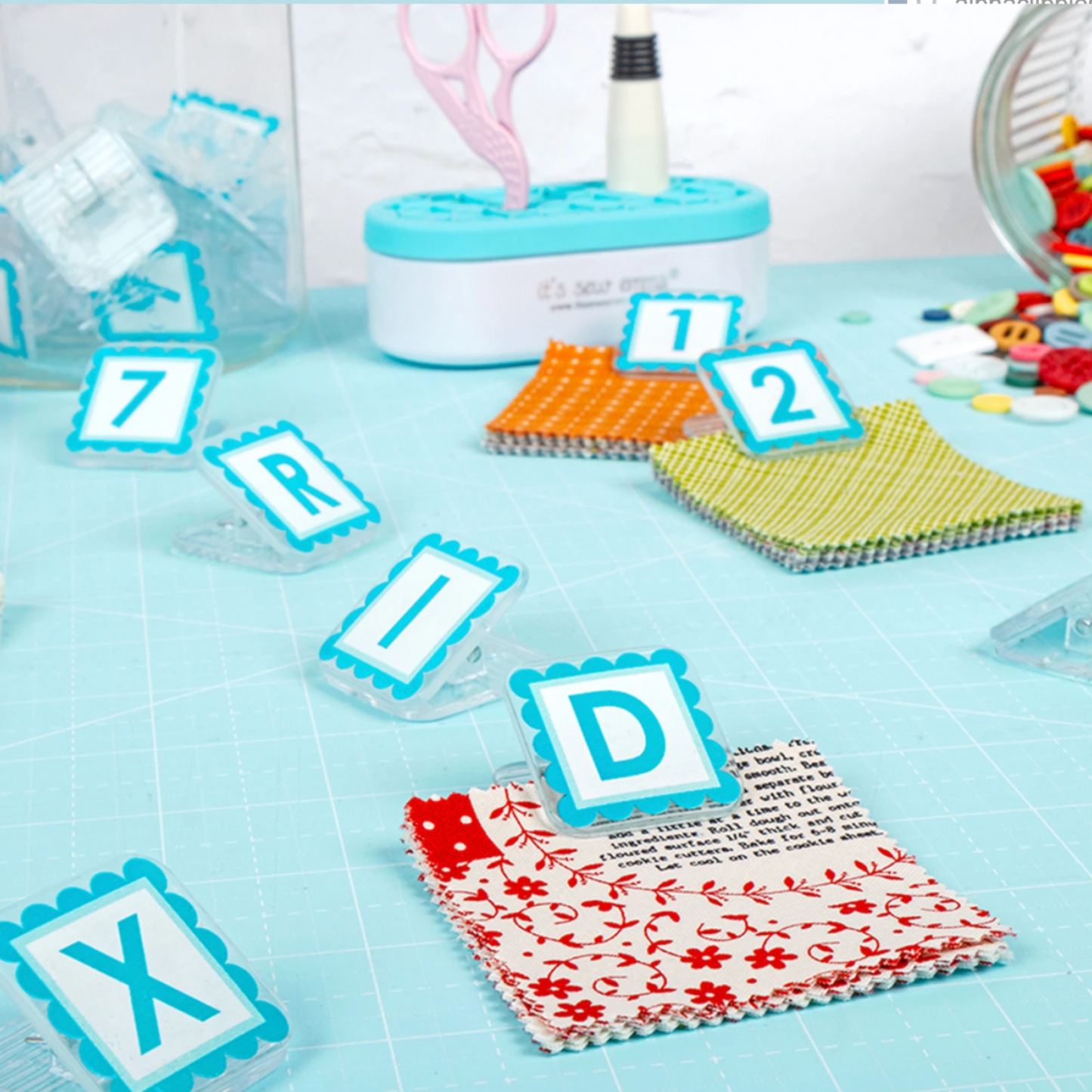 AlphaClippies quilt organization clips displayed on sewing table with fabric squares and quilting accessories
