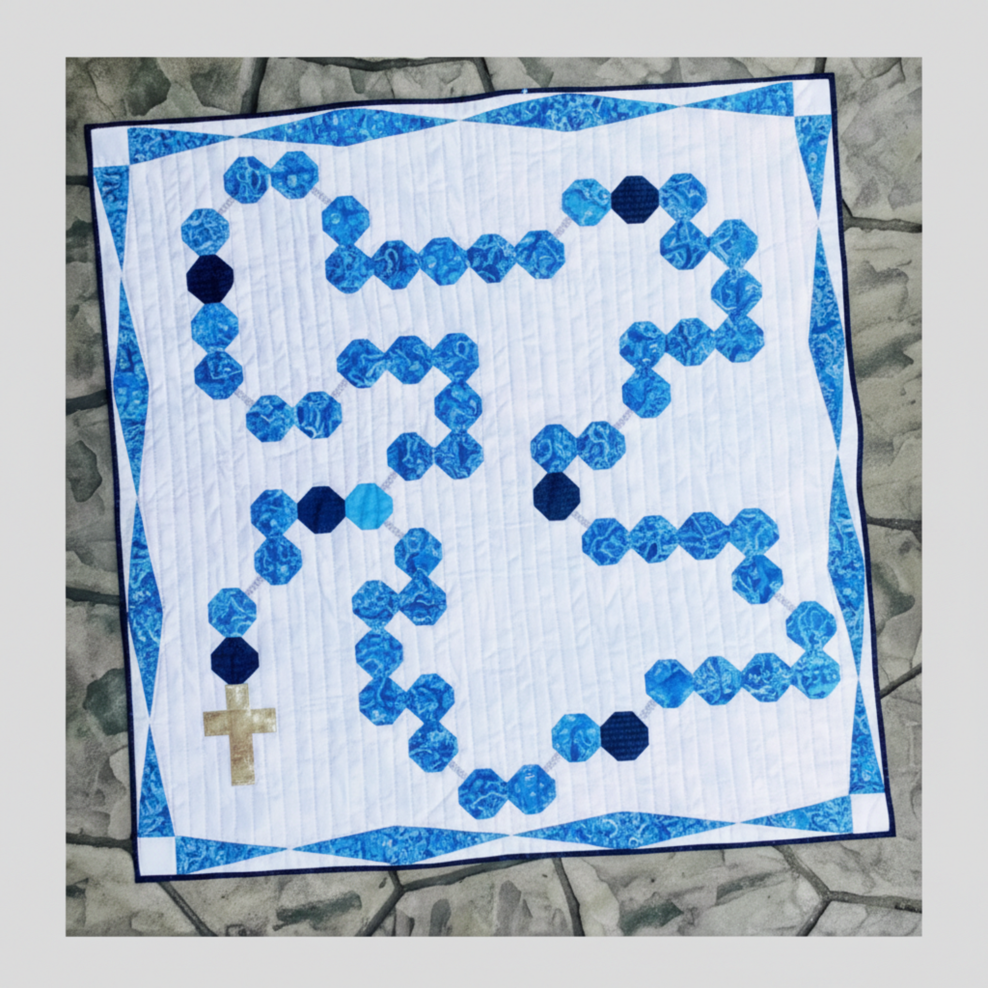 Rosary Baby Quilt pattern cover featuring blue rosary bead design quilt with white background and blue border on stone-look backdrop