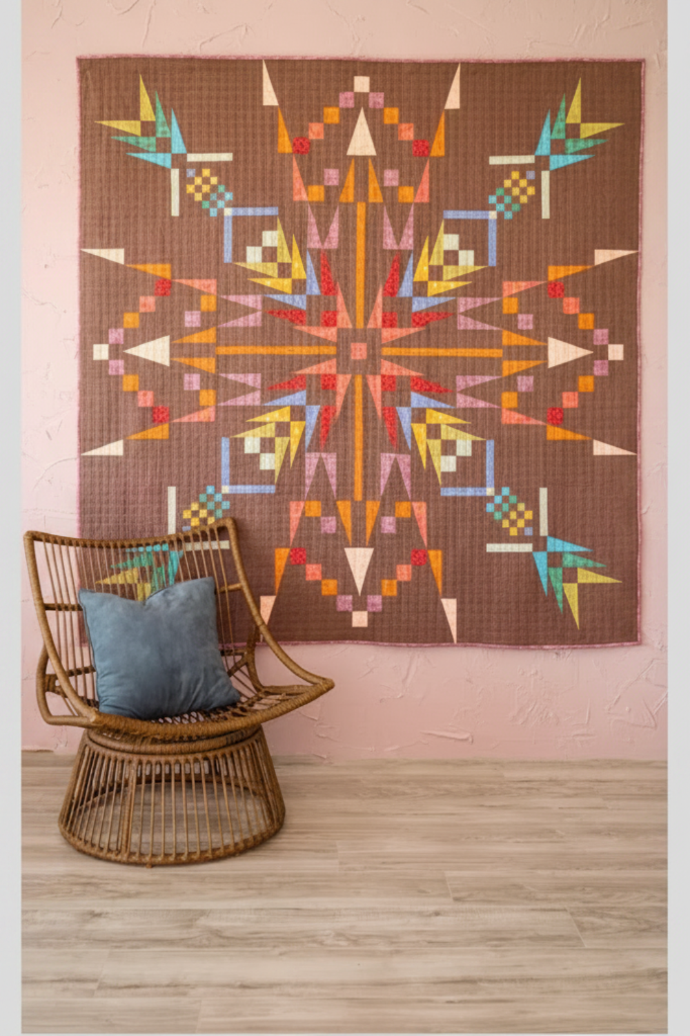Northern Lights quilt displayed on blush pink wall with colorful modern geometric design and wicker accent chair