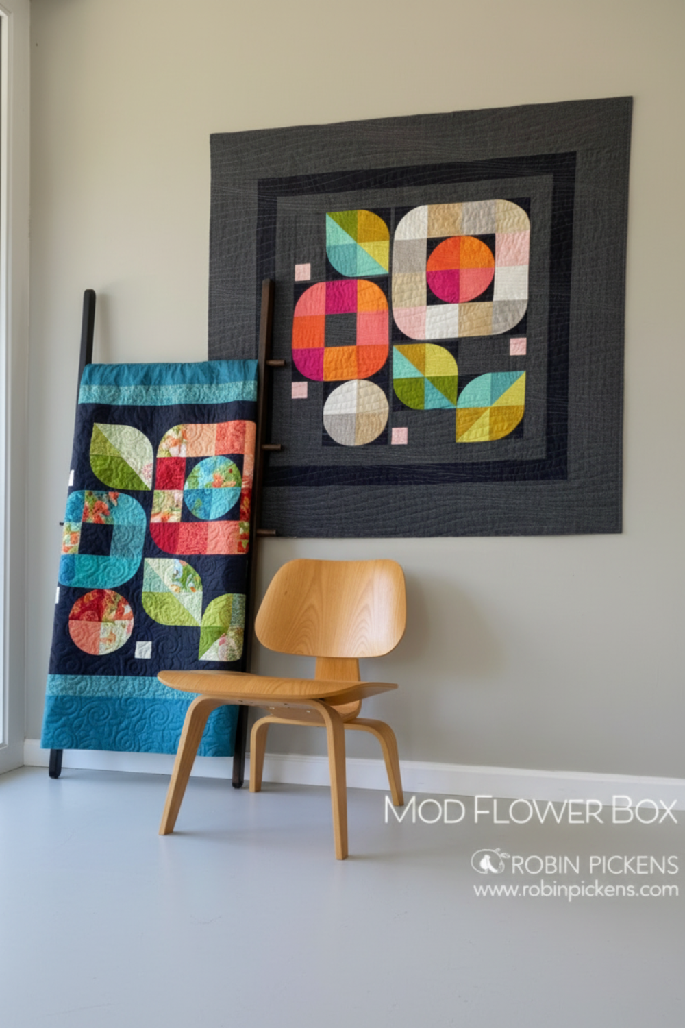 Colorful geometric quilt on a wall with a wooden chair below and a framed quilt in the background.