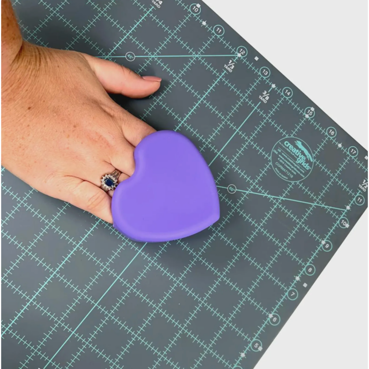 Hand holding the back side of The Gypsy Quilter heart-shaped cutting mat cleaning pad, showing ergonomic size and shape.