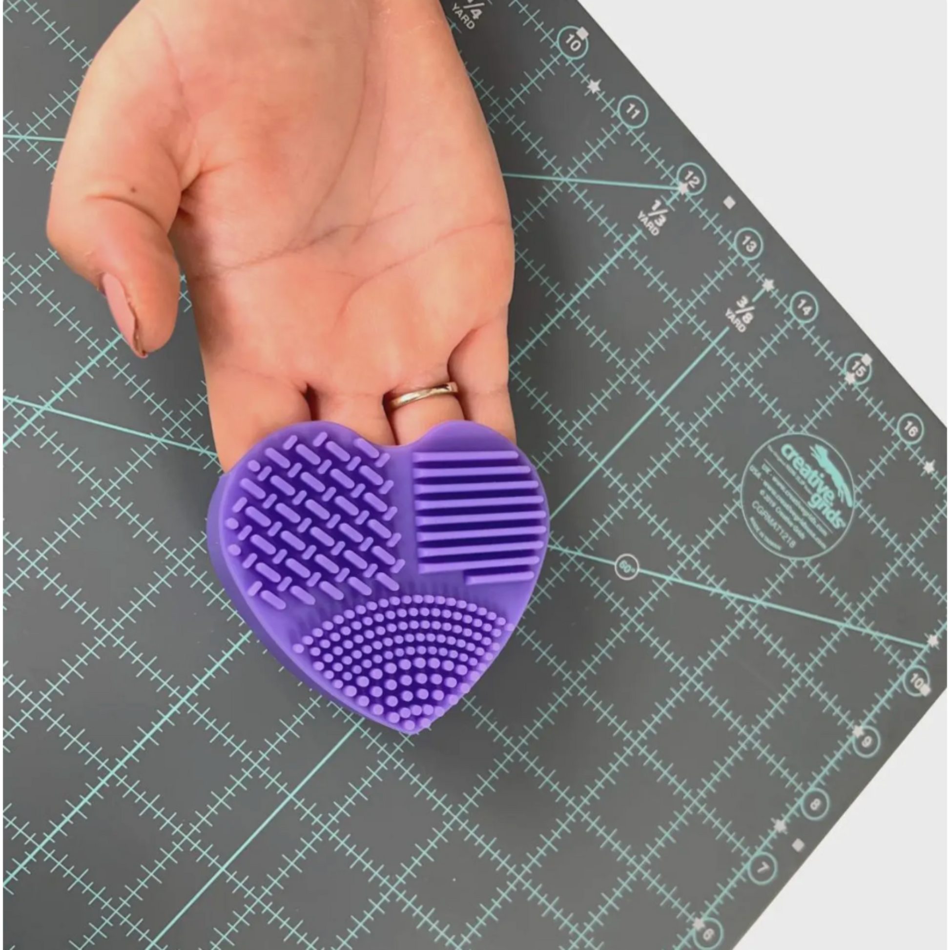 Hand holding The Gypsy Quilter heart-shaped mat cleaning pad, displaying textured silicone surface used to clean cutting mats.