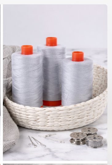 Three Aurifil Mako Cotton Thread 50wt Aluminum spools displayed in a woven basket with sewing accessories.