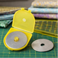 Open OLFA rotary blade case showing installed blade and extra 45mm blade on cutting mat with quilting ruler and fabric