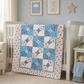 Blue and white floral baby quilt displayed on white crib in soft pastel nursery with teddy bear and nursery décor