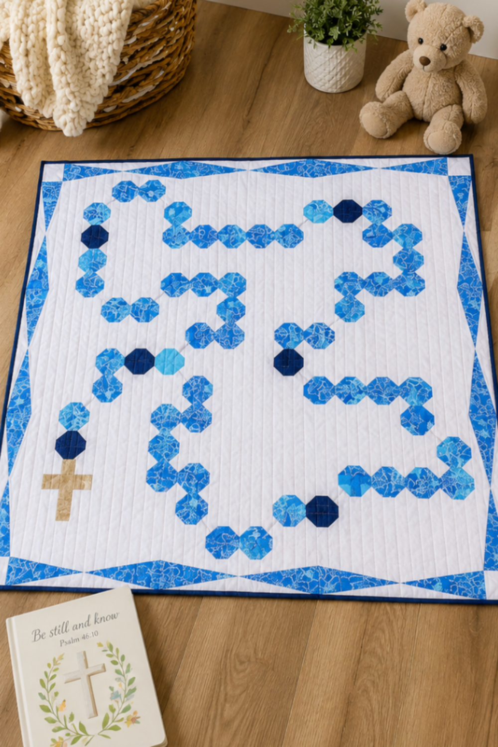 Rosary-inspired baby quilt displayed on nursery floor with teddy bear, woven basket, and inspirational Christian book