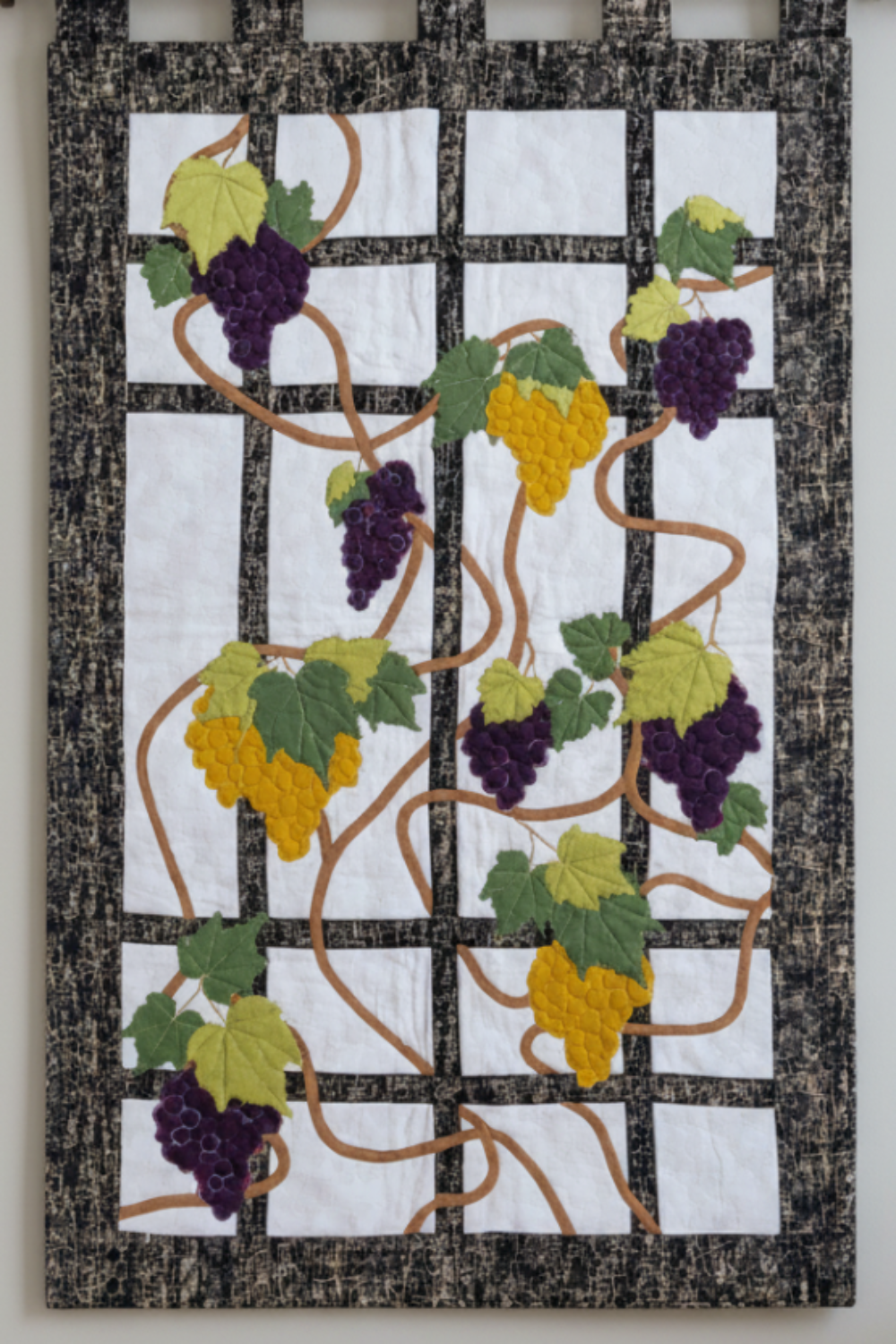Grapevine quilted wall hanging with purple and golden grapes, green leaves, and black trellis border on a clean light background.