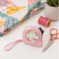 Riley Blake Quilty pink tape measure displayed beside a handmade quilt, thread spool, and embroidery scissors in a quilting workspace.