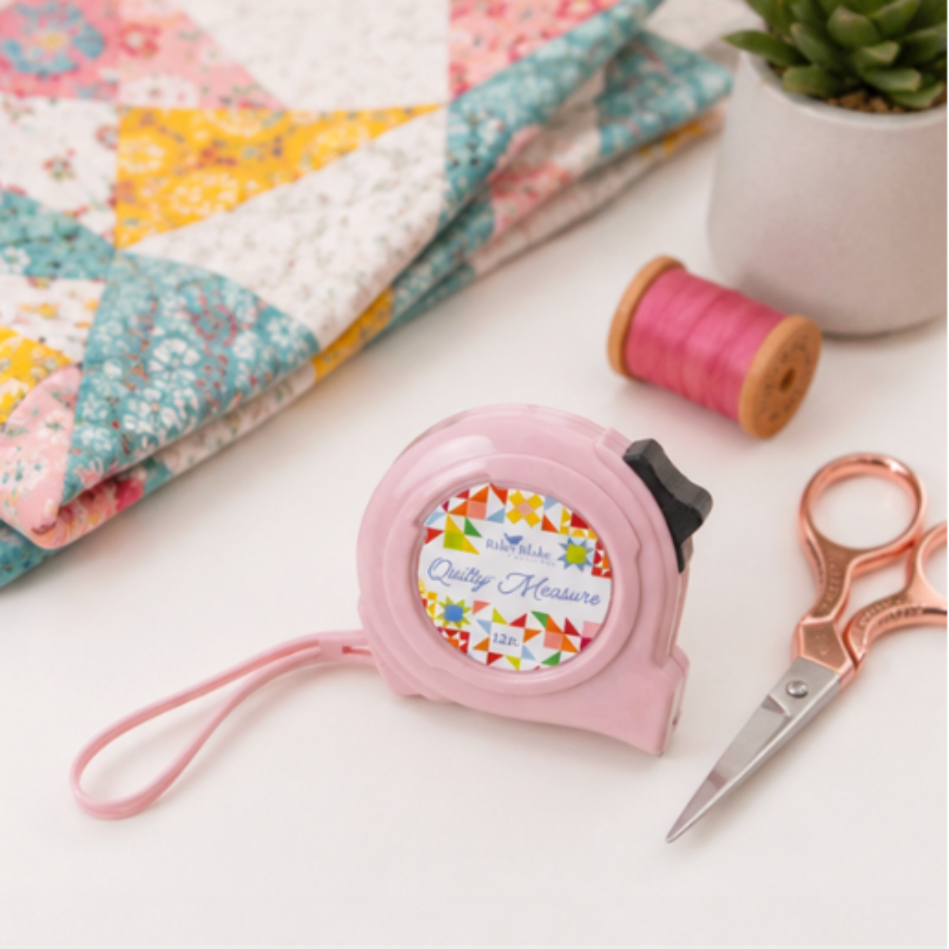 Riley Blake Quilty pink tape measure displayed beside a handmade quilt, thread spool, and embroidery scissors in a quilting workspace.