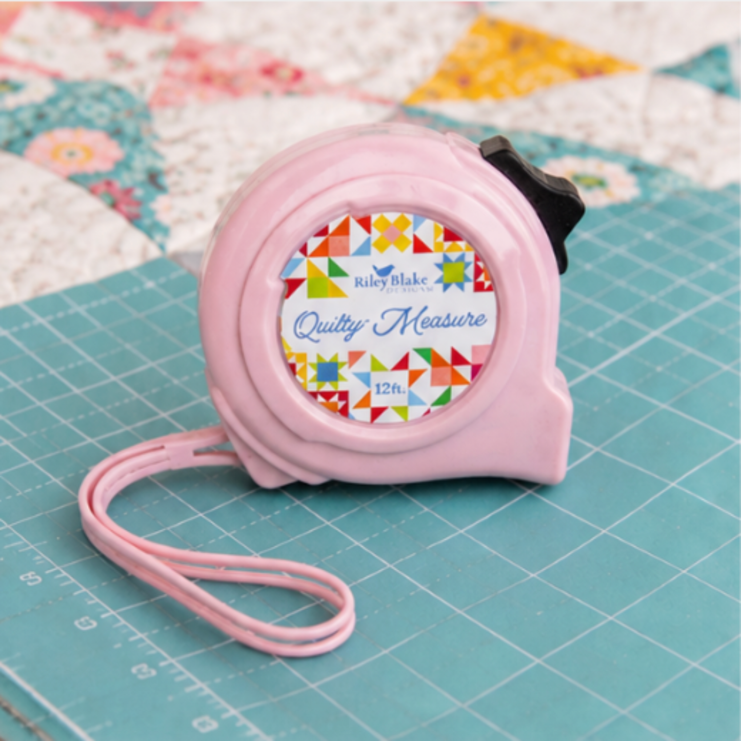 Riley Blake Quilty 12 ft pink tape measure on a quilting cutting mat with colorful patchwork quilt in the background.