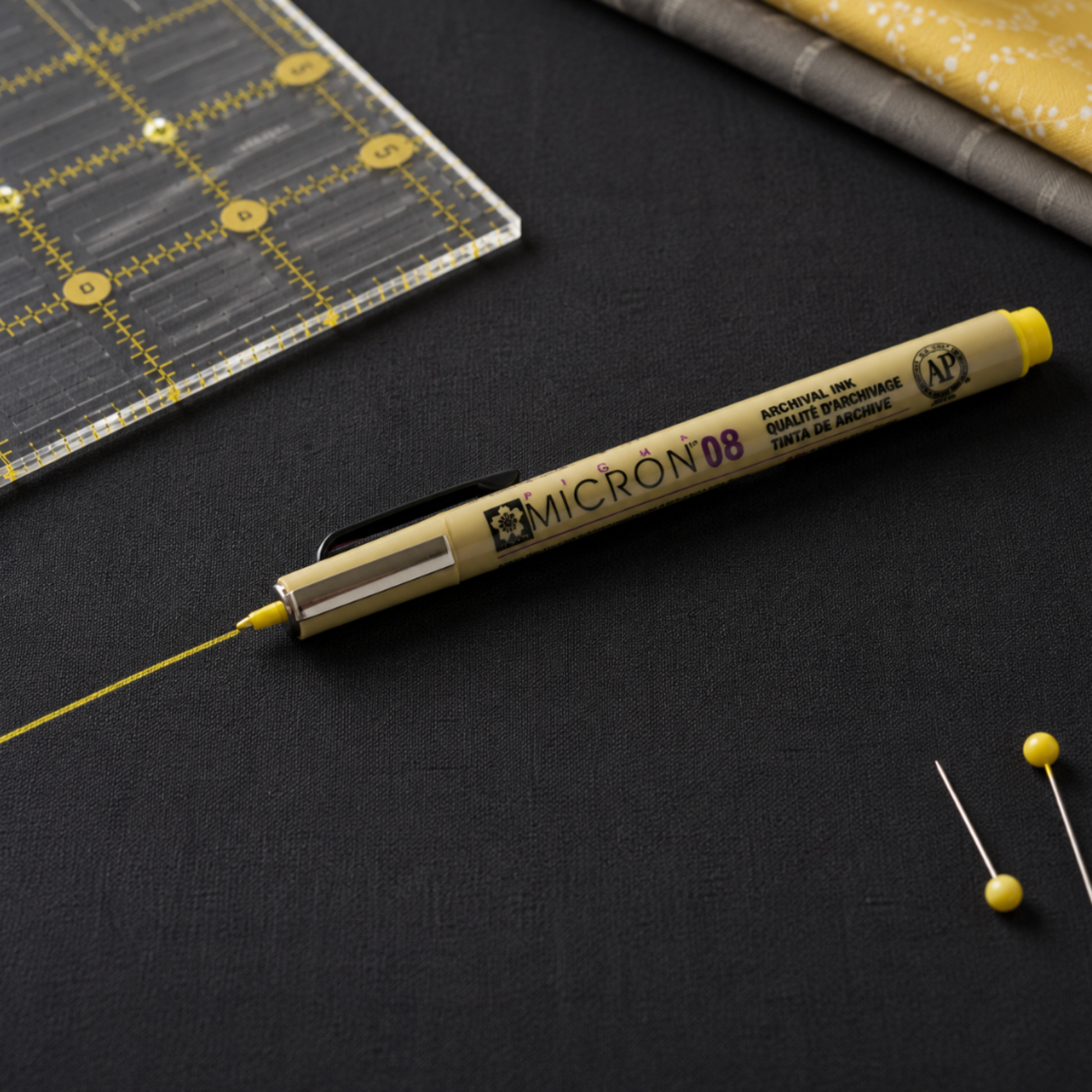 Yellow Pigma Micron 08 fabric marking pen drawing a fine yellow line on dark fabric beside a quilting ruler and sewing pins, ideal for quilting and sewing projects.