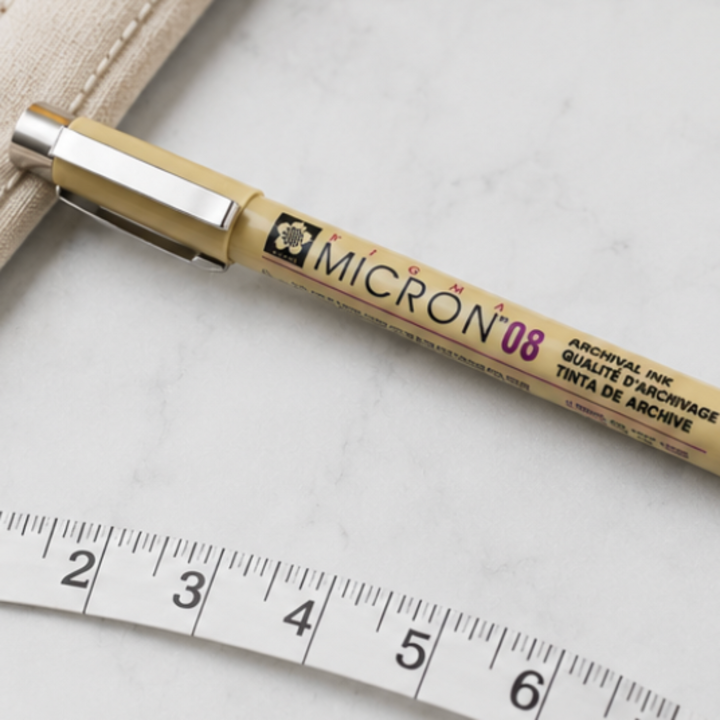Pigma Micron 08 archival ink pen displayed on a light gray background with a close-up styled flat lay featuring a measuring tape, thread spool, and sewing supplies.