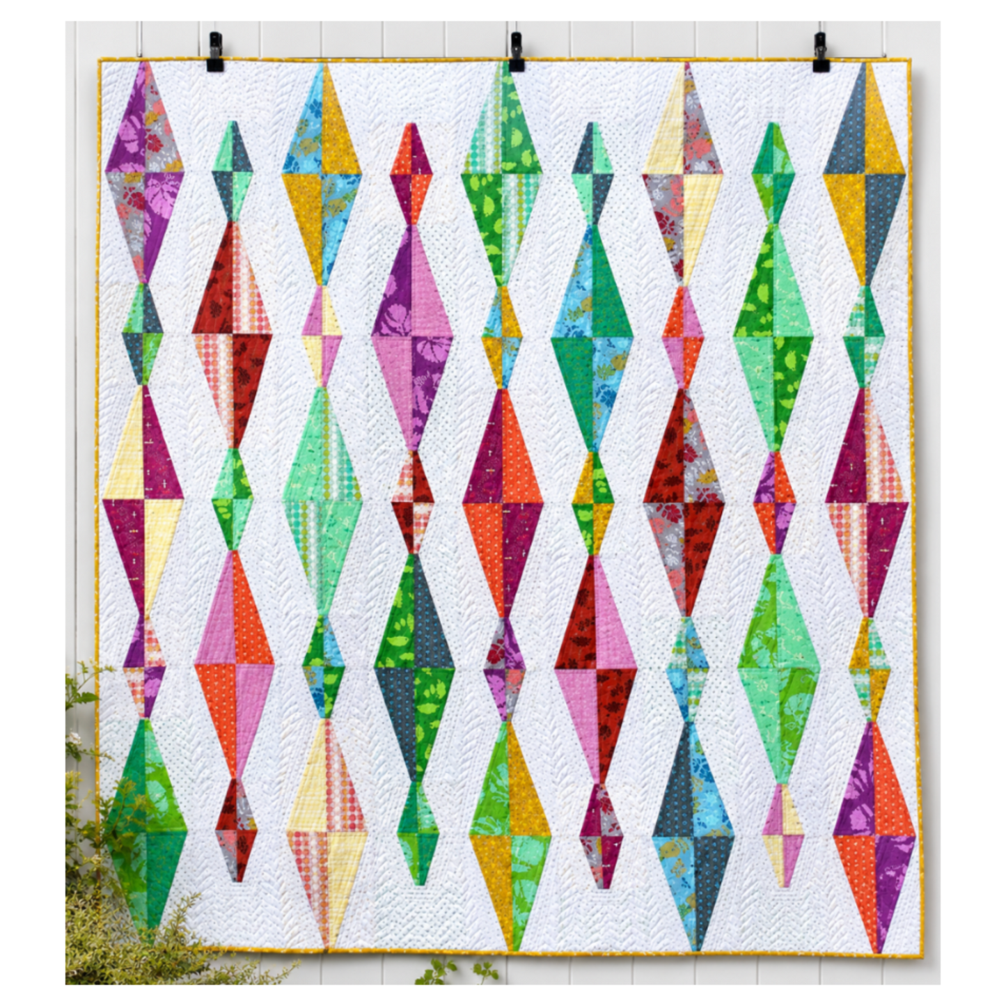 Modern Diamond Daze quilt with colorful geometric diamond blocks on a white background, quilted with flowing texture and finished with a yellow binding, displayed hanging on a white wall.