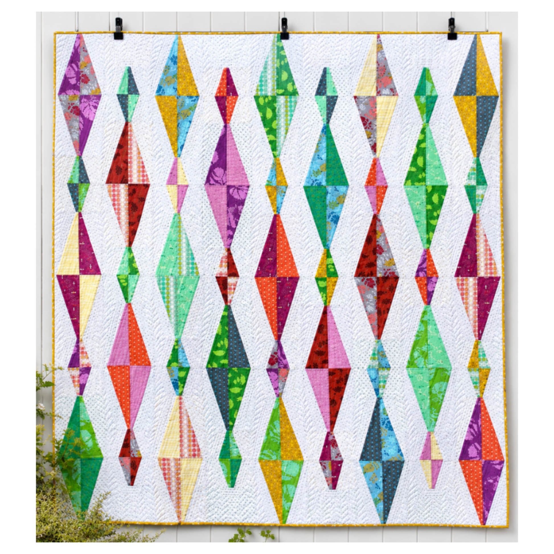 Modern Diamond Daze quilt with colorful geometric diamond blocks on a white background, quilted with flowing texture and finished with a yellow binding, displayed hanging on a white wall.