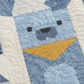 Close-up of a blue patchwork puppy quilt showing detailed quilting stitches, geometric dog face design, and yellow bandana accent on soft white fabric.
