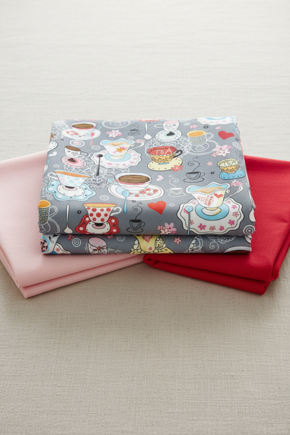 Alice Multi Teacups fabric by Jason Yenter for In The Beginning Fabrics folded on a light linen surface with coordinating pink and red fabrics – whimsical Mad Hatter tea party scatter print with teacups, saucers, hearts, and lace on soft grey, perfect for children's quilts and novelty projects, 100% cotton, 3 yard cut