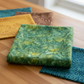 Expressions Batiks Hand-Dyes Amazon fabric by Riley Blake Designs folded on a warm wood surface with coordinating golden yellow, earthy brown, and deep teal batik swatches nearby – rich jungle green hand-dyed batik with golden-yellow undertones, perfect for nature-inspired and jewel-tone quilts, 100% cotton batik