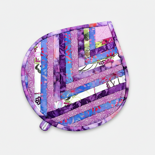  "Leaf-Shaped Quilted Hot Pad – Purple Garden, slightly angled editorial view on light grey background with soft natural shadow and hanging loop visible, showing herringbone chevron strip piecing in purple, lavender, blue, pink, magenta, and floral botanical prints with purple batik border – handmade by IraRobi Quilts