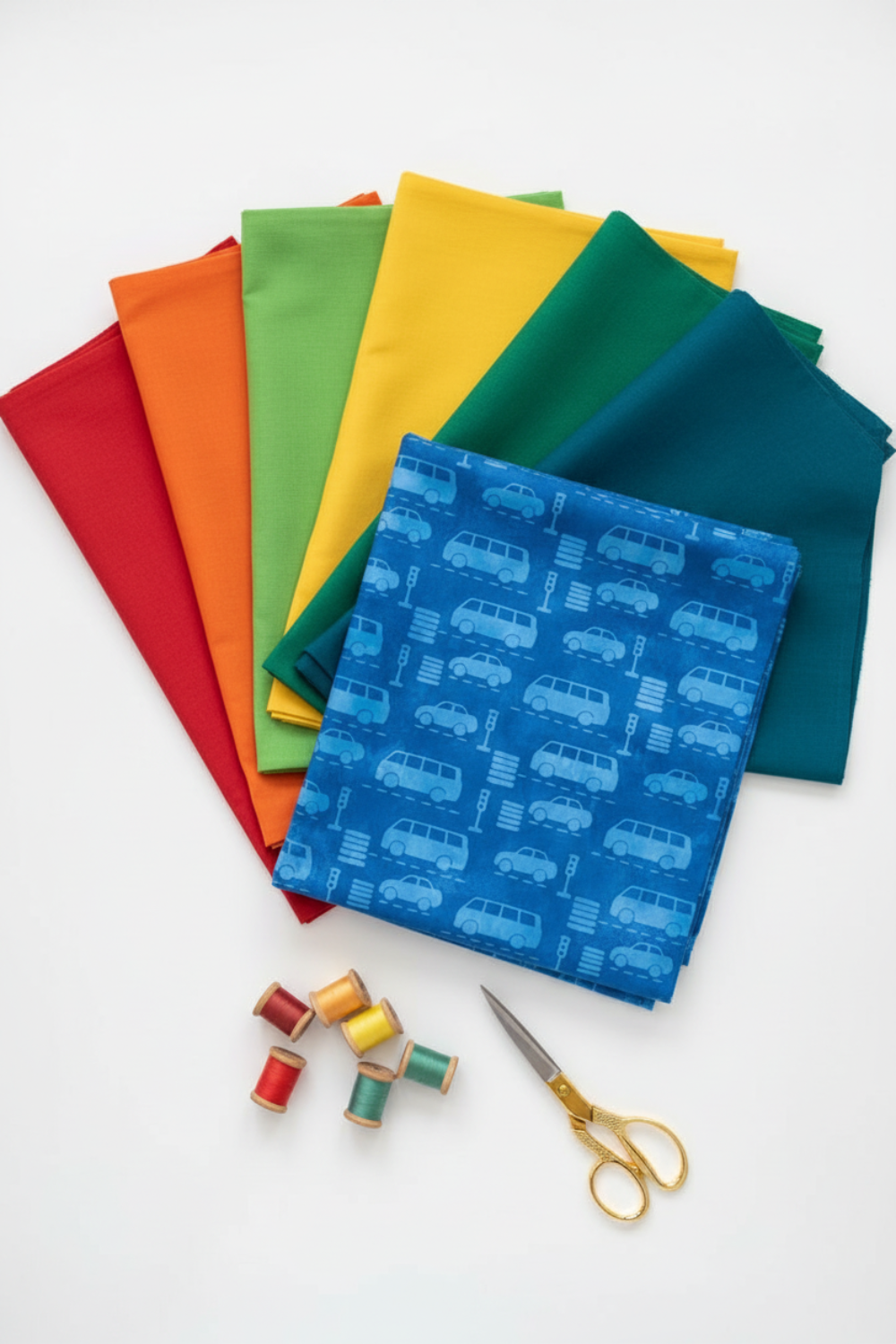 On the Go Automobiles Blue quilting cotton by Jason Yenter for In The Beginning Fabrics — vertical lifestyle fan spread with the cobalt-blue automobile print as the front hero fabric and coordinating solid-color fabrics in red, orange, yellow, green, and teal fanned behind, with thread spools and gold scissors below. Coordinating fabrics and quilt patterns available at IraRobi Quilts.
