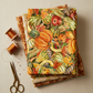 Autumn Celebration Multi Vegetables quilting cotton — vertical flat lay folded as hero on a tall coordinating autumn fabric stack with brass scissors and amber thread spools on a warm cream linen surface. Coordinating fabrics and quilt patterns available at IraRobi Quilts.