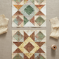 Quilting work-in-progress flat lay showing two Barn Star quilt blocks with Expressions Batiks Breathe Ecru fabric by Riley Blake Designs as the cream background – warm cream and ecru tonal batik with pine needle and feathery frond motif in soft taupe and golden-beige, with coordinating sage green, warm taupe, and muted terracotta tonal batik accent pieces, loose cut fabric pieces and cream thread spool on a warm linen surface