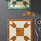 Bear's Paw quilt block work-in-progress featuring Expressions Batiks Hand-Dyes Orange Multi I fabric by Riley Blake Designs as the center square – complex multicolor hand-dyed batik with warm golden amber-orange, olive green, slate blue-grey, and burnt red-orange, with warm brown tonal batik border and accent triangles on cream background, loose cut fabric pieces, tape measure, and scissors on dark grey concrete surface