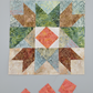 Bear's Paw quilt block work-in-progress close-up featuring Expressions Batiks Hand-Dyes Sunset fabric by Riley Blake Designs as the center diamond – soft coral-pink hand-dyed batik with warm golden-orange splashes, soft peach tones, and hints of deeper rose, with light blue-grey dotted batik background, warm tan-brown and forest green swirl batik triangles, and medium brown patterned batik corners, loose cut Sunset fabric squares at bottom edge, on light grey surface. Quilt blocks shown for inspiration. Coo