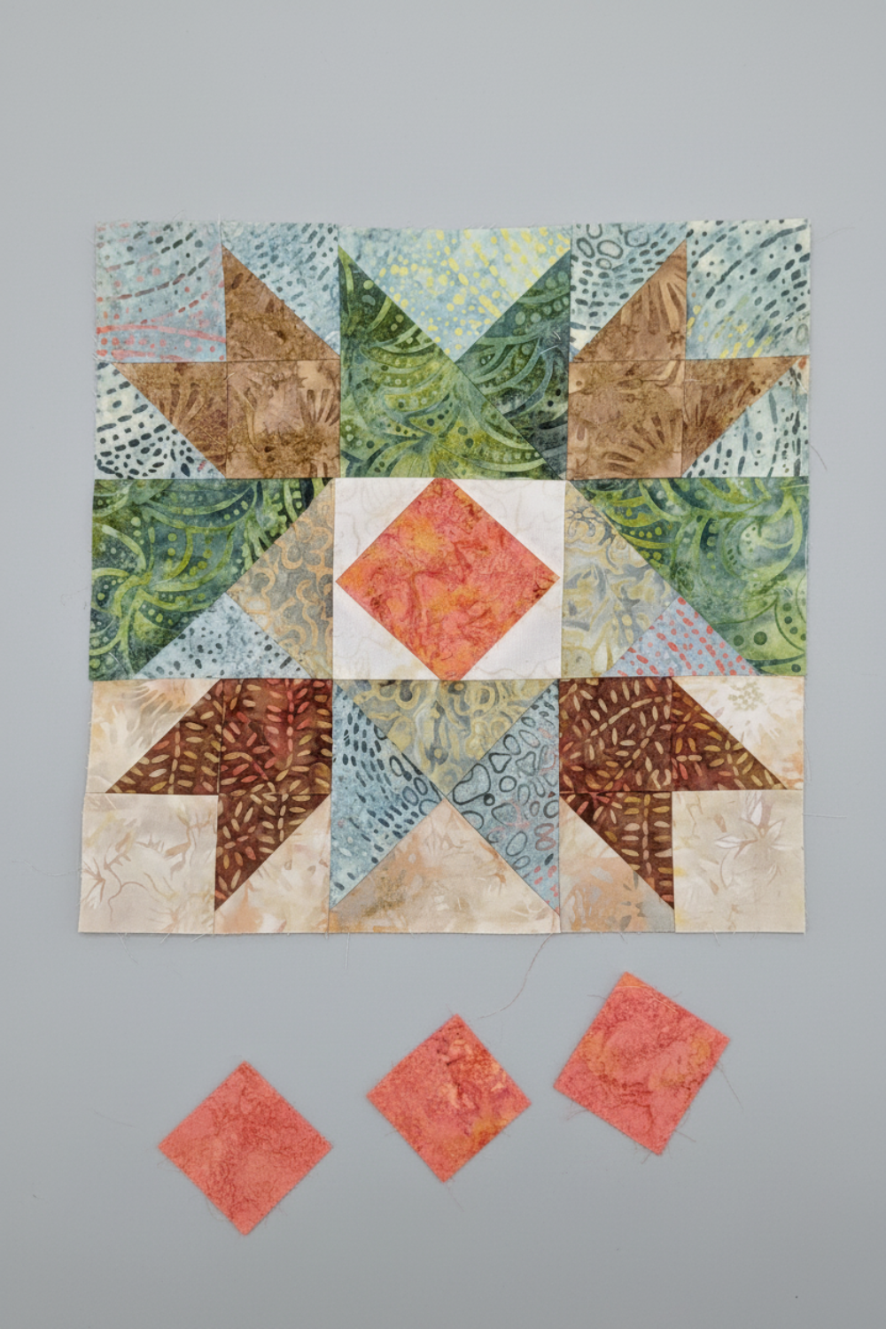 Bear's Paw quilt block work-in-progress close-up featuring Expressions Batiks Hand-Dyes Sunset fabric by Riley Blake Designs as the center diamond – soft coral-pink hand-dyed batik with warm golden-orange splashes, soft peach tones, and hints of deeper rose, with light blue-grey dotted batik background, warm tan-brown and forest green swirl batik triangles, and medium brown patterned batik corners, loose cut Sunset fabric squares at bottom edge, on light grey surface. Quilt blocks shown for inspiration. Coo