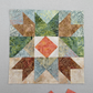 Bear's Paw quilt block work-in-progress close-up featuring Expressions Batiks Hand-Dyes Sunset fabric by Riley Blake Designs as the center diamond – soft coral-pink hand-dyed batik with warm golden-orange splashes, soft peach tones, and hints of deeper rose, with light blue-grey dotted batik background, warm tan-brown and forest green swirl batik triangles, and medium brown patterned batik corners, loose cut Sunset fabric squares at bottom edge, on light grey surface. Quilt blocks shown for inspiration. Coo