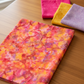 Expressions Batiks Hand-Dyes Bouquet fabric by Riley Blake Designs folded on a warm wood surface with coordinating hot pink, golden yellow, and soft lavender tonal batik swatches nearby – vibrant multicolor hand-dyed batik with golden orange base and bold splashes of hot pink, magenta, crimson, and lavender, perfect for floral and scrappy quilts, 100% cotton batik