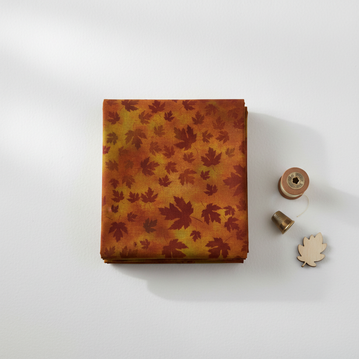 Autumn Celebration Brown Maple Leaves tonal blender quilting cotton by Jason Yenter for In The Beginning Fabrics — neatly folded fabric stack showing tossed dark burgundy-maroon maple leaf silhouettes on mottled burnt amber and rust watercolor ground, styled with a wooden thread spool, thimble, and leaf accent on a white surface. 100% cotton, 43–44" wide.