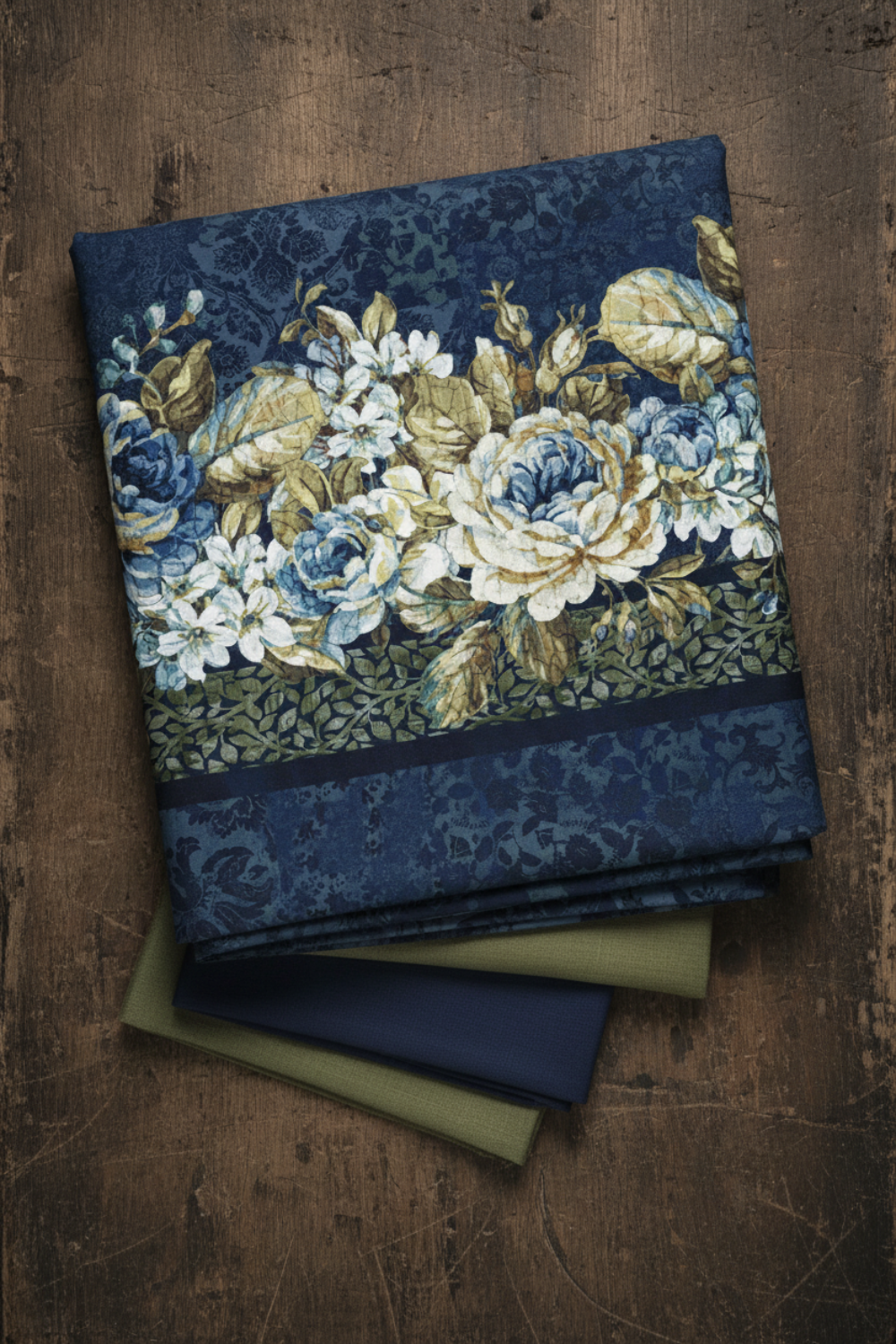 Carpet Bag Romance Blue Border Print fabric by Jason Yenter for In The Beginning Fabrics draped on a dark wood surface – lush cascading floral garland stripe with large ivory and blue roses, white hydrangeas, and olive foliage on deep navy damask ground, perfect for fussy-cut borders and vintage quilt projects, 100% cotton, 3 yard cut