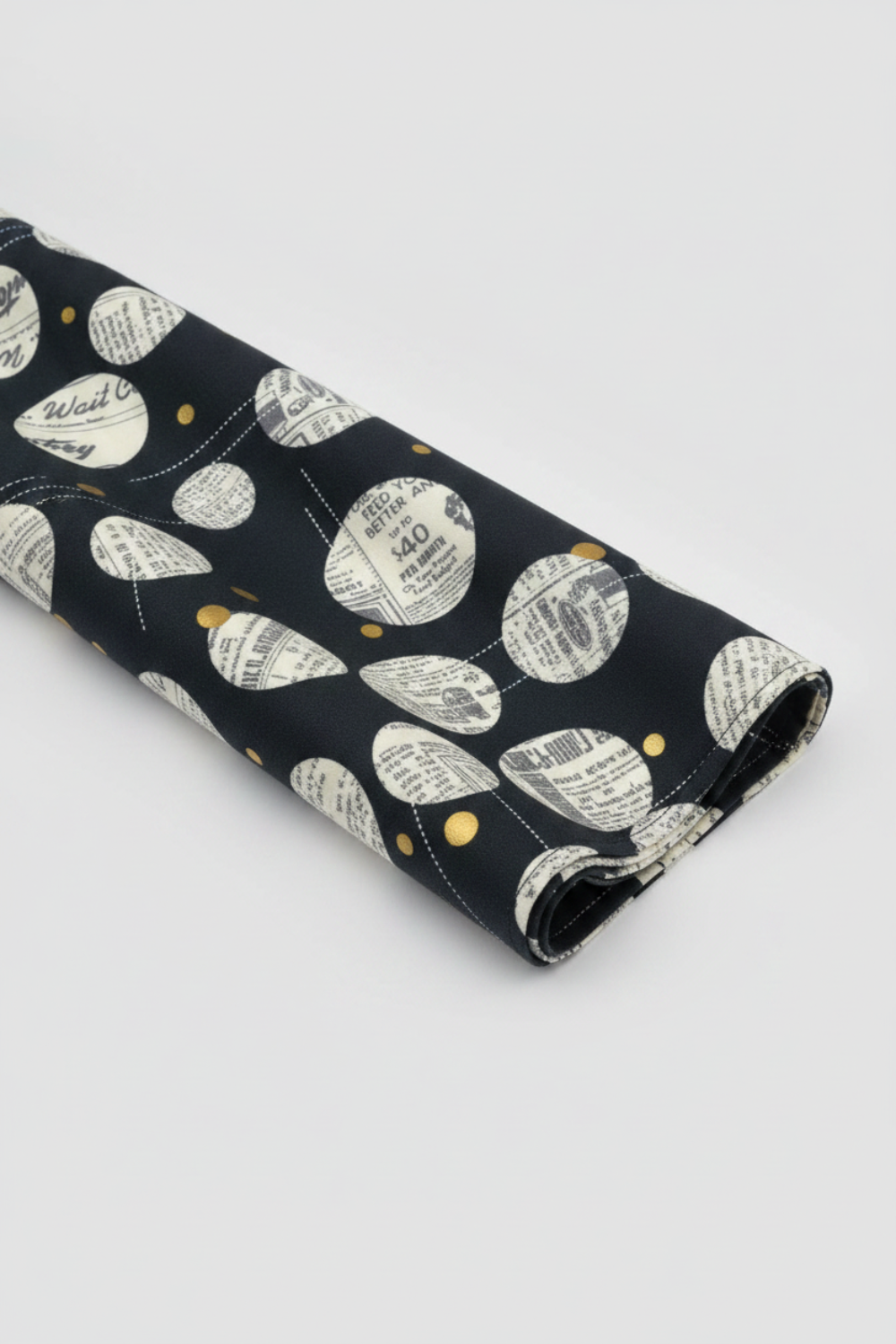 Celestial News Dropping Onyx by Zen Chic for Moda Fabrics — bold black cotton with cream newspaper circles and gold dots, folded in a diagonal sweep showing fabric drape and body. 