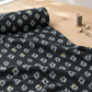 Dance in Paris Lanterns Metallic Charcoal fabric unrolled across a light wood sewing table, showing pale gray diamond motifs and scattered gold accents beside a thread spool and fabric clips.