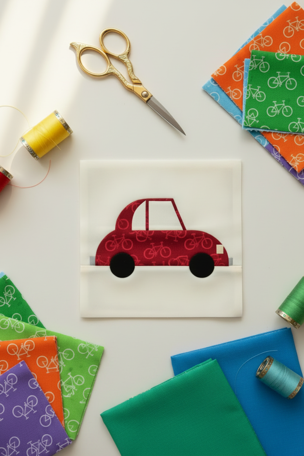 On the Go Bicycles Red quilting cotton by Jason Yenter for In The Beginning Fabrics — vertical lifestyle flat lay of a Cool Cars quilt block by Sew Kind of Wonderful made with the Quick Curve Ruler, featuring the red bicycle tone-on-tone print as the car body, with solid rainbow fabric folds, colorful thread spools, and gold scissors on a white surface. Quilts shown for inspiration. Coordinating fabrics and quilt patterns are available at IraRobi Quilts.