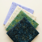 Four folded Riley Blake Designs fabrics arranged in a fan: Color Play Mottled Rainstorm, Expressions Batiks Amazon, Breathe Laurel, and Periwinkle, in dark teal, green, cream, and lavender.