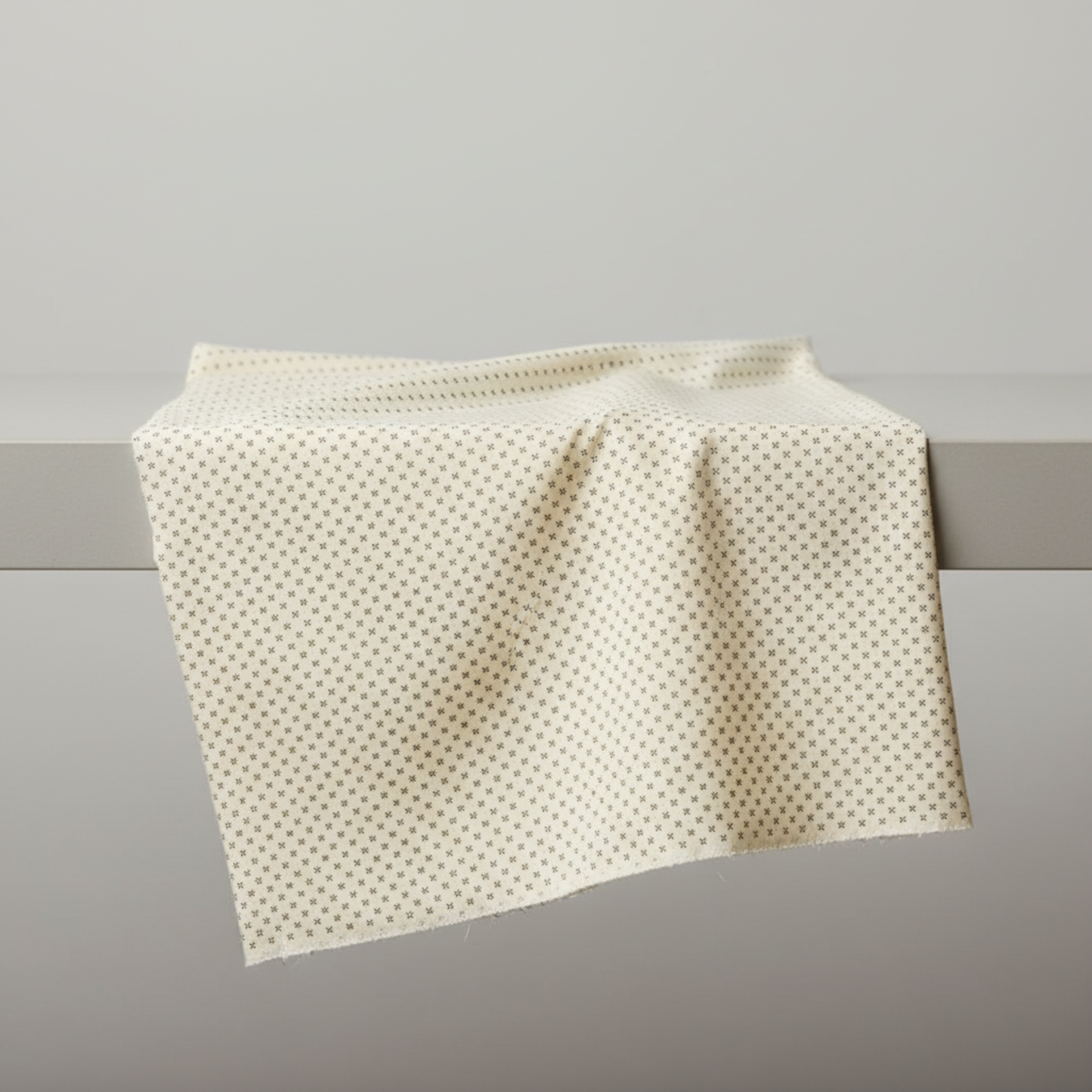 Scraps of Kindness Cream Nine Patch Boxes quilting cotton by Kim Diehl for Henry Glass — fabric draped over a grey surface showing natural folds and raw cut edge, warm cream ground with evenly spaced tiny dark brown four-point cross clusters