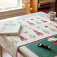 Winter Solstice II Cream Snow Swirl Fabric by Jason Yenter for In The Beginning Fabrics — shown as the background and sashing fabric in the Snowy Pines Table Runner by Margot Languedoc, cozy sewing room work in progress with fairy lights and a mug of tea. Quilts shown for inspiration feature the Snowy Pines Table Runner by Margot Languedoc for The Pattern Basket — available at IraRobi Quilts.