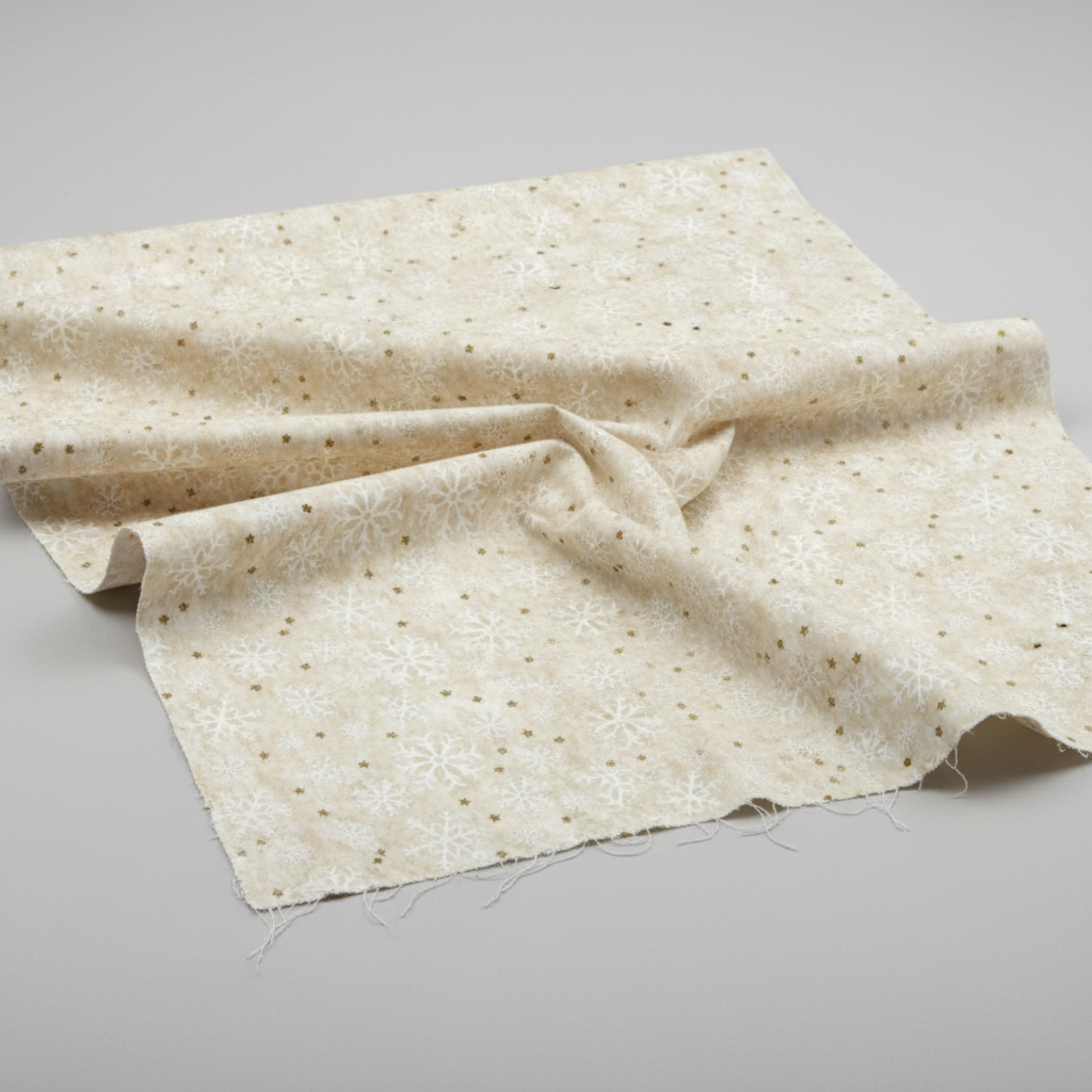 Winter Solstice II Cream Snowflakes Metallic quilting cotton by Jason Yenter for In The Beginning Fabrics — fabric draped over a grey surface showing natural folds and raw cut edge, tossed white snowflakes with gold metallic star dots on warm mottled ecru and cream ground. Coordinating fabrics and quilt patterns are available at IraRobi Quilts.