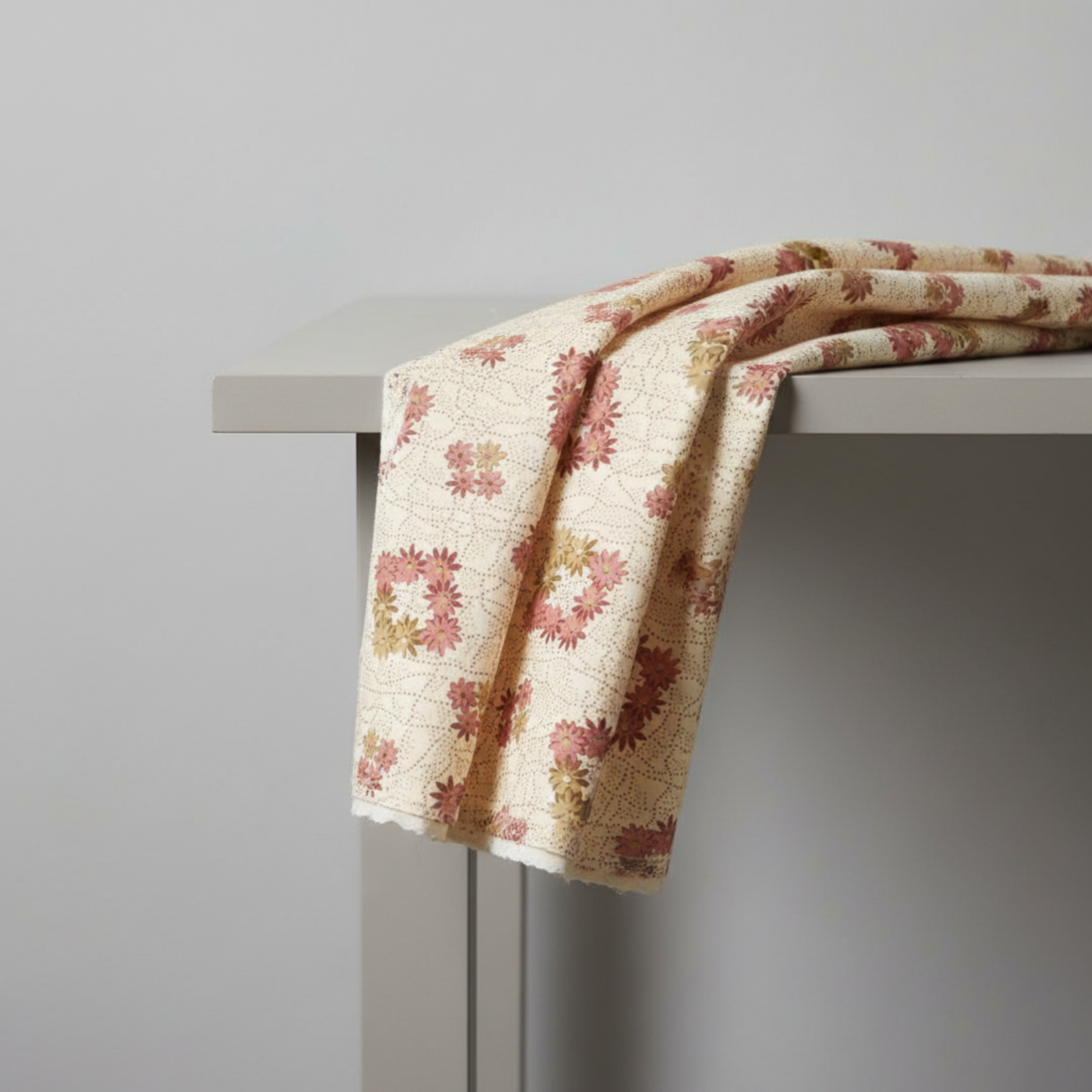 The Fox Homestead Cream Square Wreath quilting cotton by Stacy West for Henry Glass — fabric draped over a grey surface showing natural folds and raw cut edge, cream ground with tossed daisy wreath clusters in dusty rose, burgundy, and olive