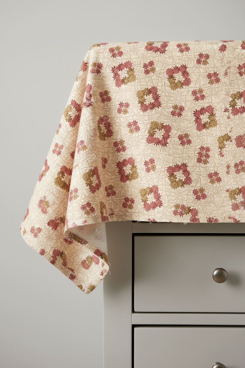 The Fox Homestead Cream Square Wreath quilting cotton draped over a light grey table edge — warm cream with tossed daisy clusters in dusty rose, muted burgundy, and olive-khaki, raw cut edges, by Stacy West for Henry Glass