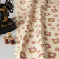 The Fox Homestead Cream Square Wreath quilting cotton by Stacy West for Henry Glass — vertical close-up showing fabric in soft folds with raw cut edge, cream ground with daisy wreath clusters in dusty rose, burgundy, and olive, with vintage sewing machine detail and thread bobbins