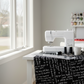 Writing on the Wall Curves Black quilting cotton by Sew Kind of Wonderful for FreeSpirit Fabrics -- bright modern sewing studio with black curves phrase fabric draped over a white sewing table near a large window, modern white sewing machine, curved acrylic ruler, thread spools, scissors, and ceramic mug in warm natural light