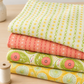 Four coordinating Dandi Duo fabrics stacked on a sewing table beside thread and scissors: cream, peach, green paisley, and yellow dandelion prints.