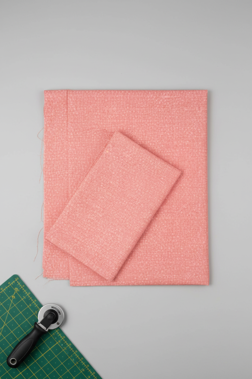 Dotty Thatched Carnation quilting cotton by Robin Pickens for Moda Fabrics — partially folded fabric on a light grey surface with a cutting mat corner and rotary cutter. Warm coral-pink ground with scattered tonal dots and crosshatch texture. Coordinating fabrics and quilt patterns available at IraRobi Quilts.