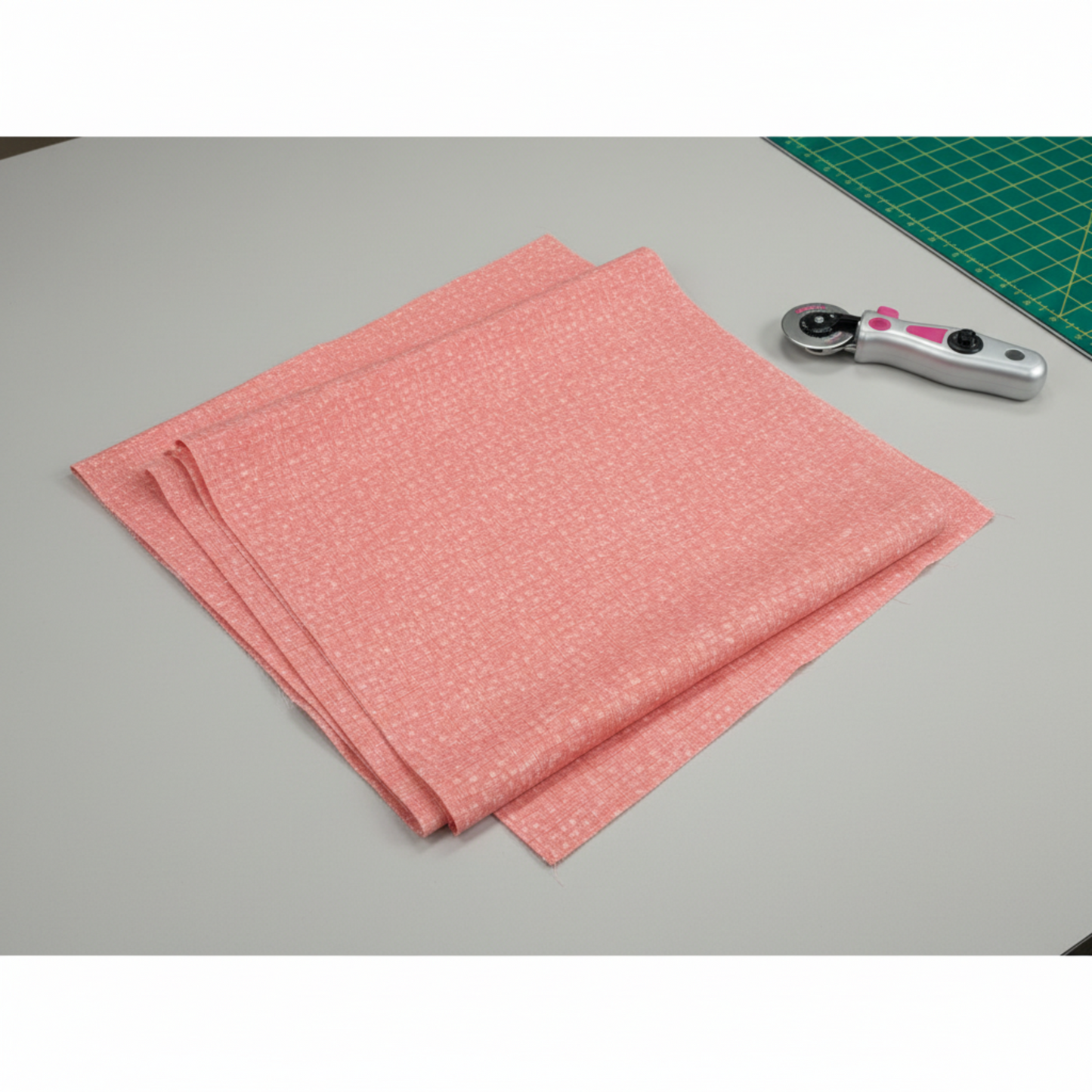 Dotty Thatched Carnation quilting cotton by Robin Pickens for Moda Fabrics — partially folded fabric on a light grey surface with a cutting mat corner and rotary cutter. Warm coral-pink ground with scattered tonal dots and crosshatch texture. Coordinating fabrics and quilt patterns available at IraRobi Quilts.