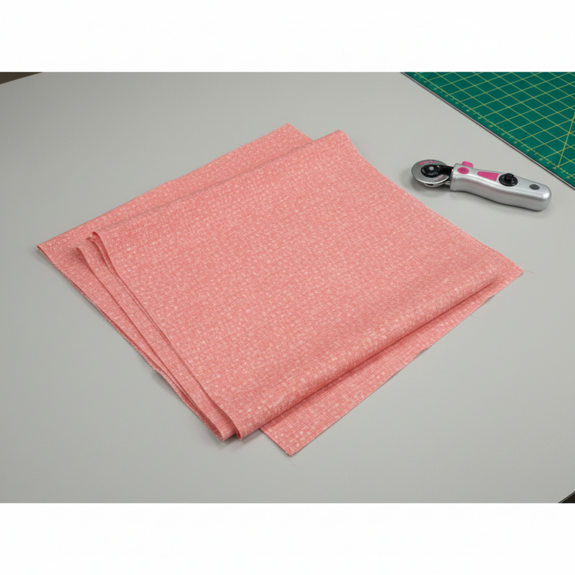 Dotty Thatched Carnation quilting cotton by Robin Pickens for Moda Fabrics — partially folded fabric on a light grey surface with a cutting mat corner and rotary cutter. Warm coral-pink ground with scattered tonal dots and crosshatch texture. Coordinating fabrics and quilt patterns available at IraRobi Quilts.
