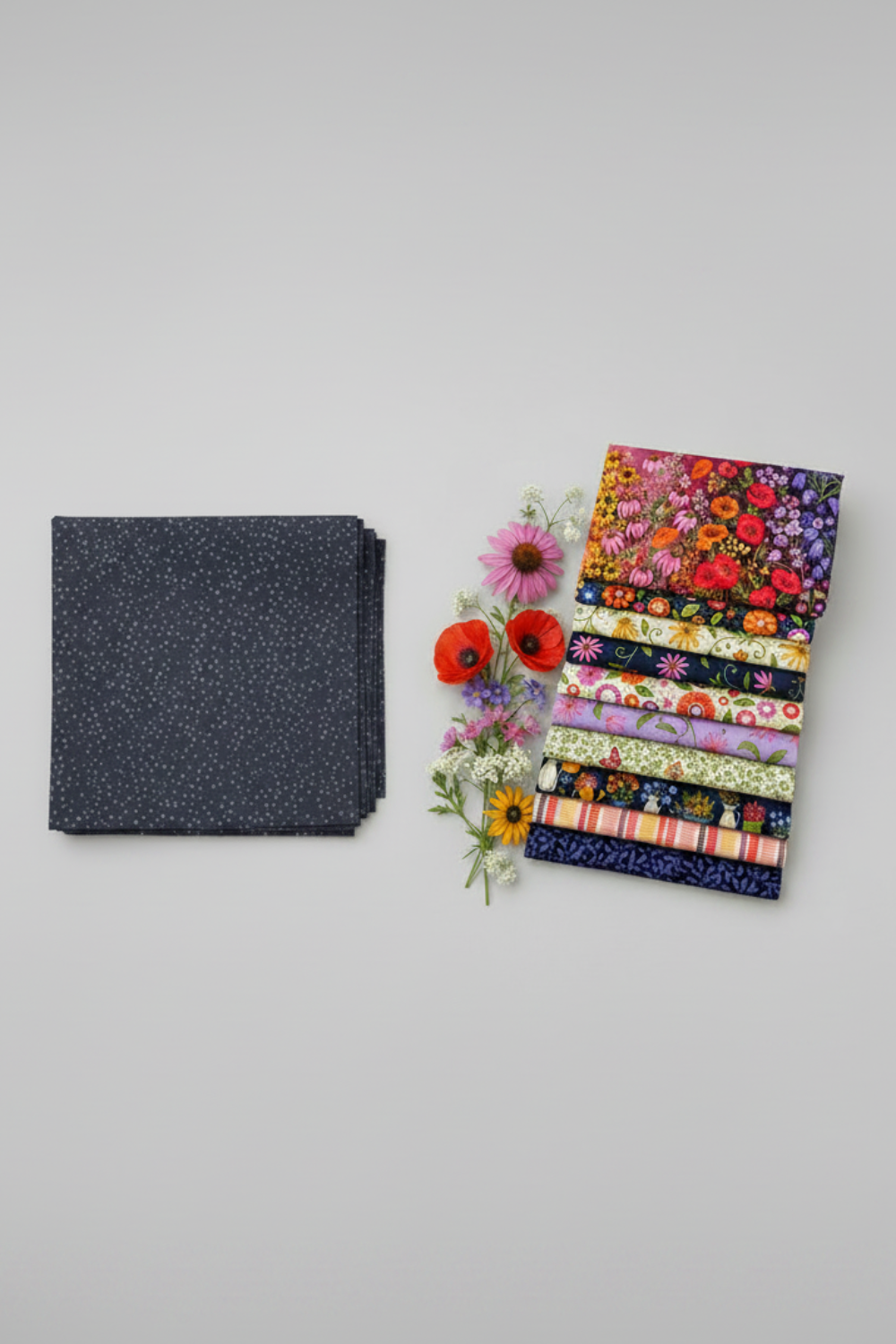 Dotty Thatched Indigo fabric by Robin Pickens for Moda Fabrics — deep indigo-charcoal with scattered tonal grey dots — shown beside a coordinating stack of Wild Blossoms quilting cotton fabrics by Robin Pickens for Moda Fabrics with fresh botanical flowers. 100% cotton, 43–44" wide. Coordinating fabrics and quilt patterns available at IraRobi Quilts.