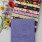 Dotty Thatched Periwinkle fabric by Robin Pickens for Moda Fabrics — periwinkle-blue with scattered tonal dots — shown beside a coordinating stack of Superbloom quilting cotton fabrics by Robin Pickens for Moda Fabrics with vibrant botanical florals. Coordinating fabrics and quilt patterns available at IraRobi Quilts.