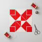Double Pinwheel quilt block layout featuring Expressions Batiks Hand-Dyes Tomato Red fabric by Riley Blake Designs – bold vivid tomato red tonal hand-dyed batik triangles and squares paired with crisp white-on-white red polka dot fabric in a dynamic pinwheel arrangement on a light grey surface, with red thread spools, silver thimble, and fabric scissors nearby. Quilt blocks shown for inspiration. Coordinating fabrics and quilt patterns are available at IraRobi Quilts.